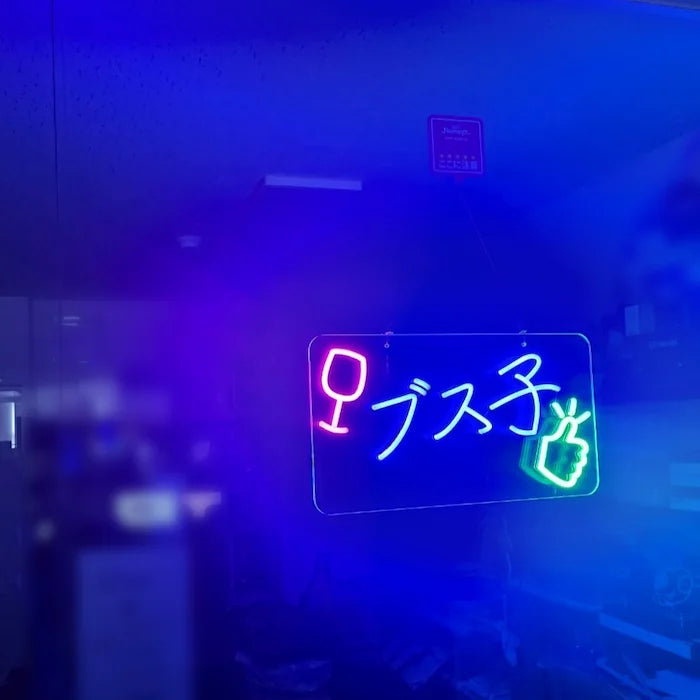 ブス子ロゴのLEDネオンサイン看板。窓ガラスタイプを薄暗い背景をバックに点灯し、左側から撮影した全体写真。