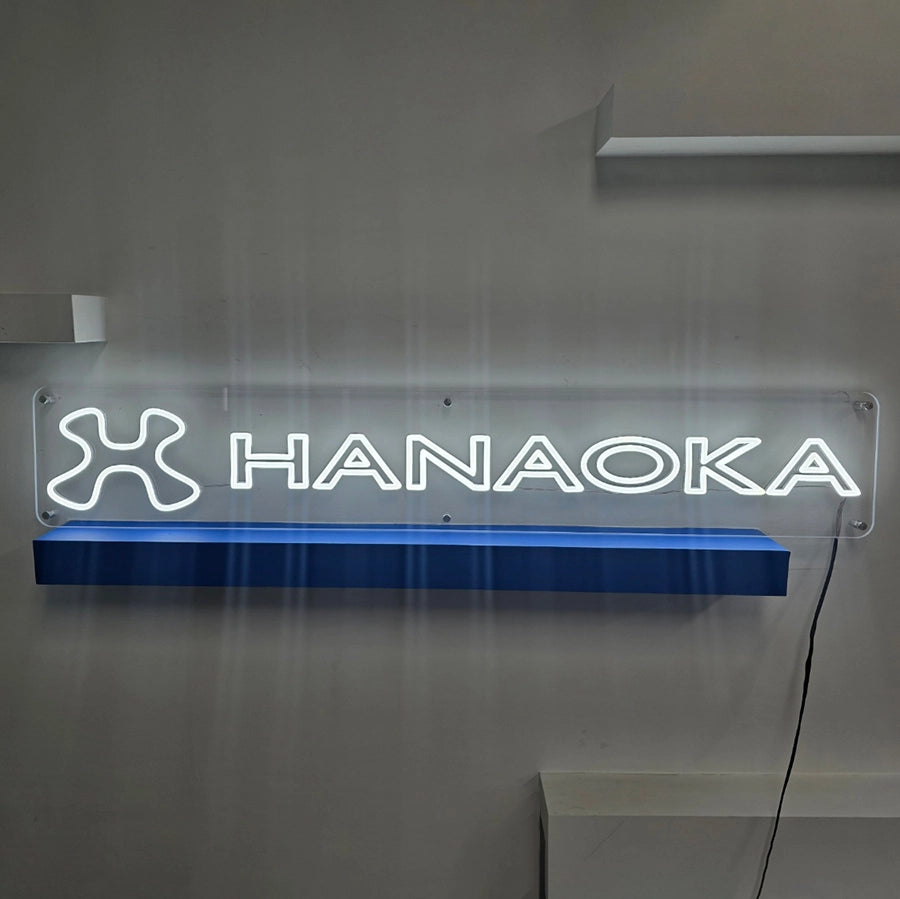 HANAOKAのシンボルマークとロゴタイプをホワイトLEDネオンで表現した企業向けオリジナルサイン製作事例。透明アクリルベースに固定し、6カ所のビスで壁面へ設置する仕様。
