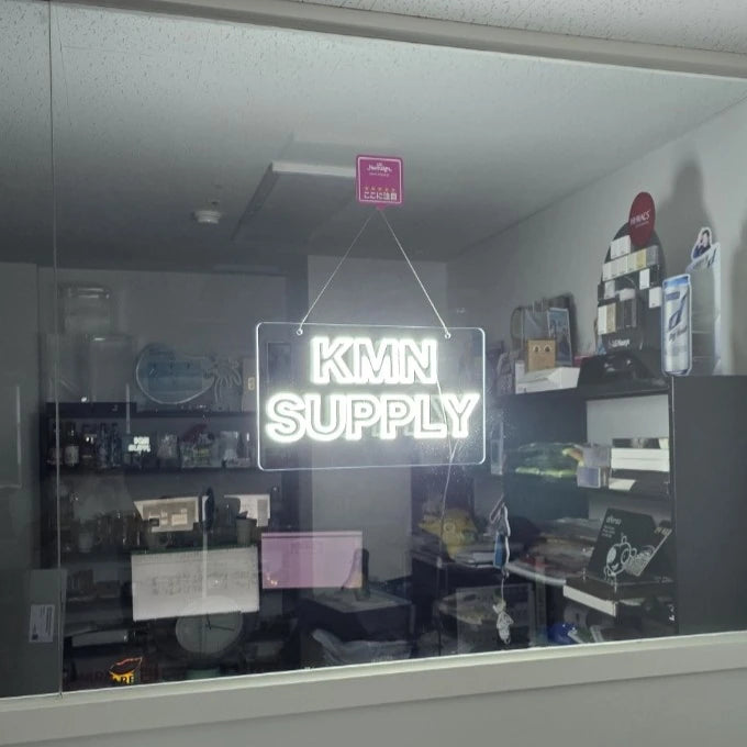 KMNSUPPLYロゴのLEDネオンサイン看板。暗い室内で窓ガラスタイプをガラスに設置して左斜めから見た全体写真。
