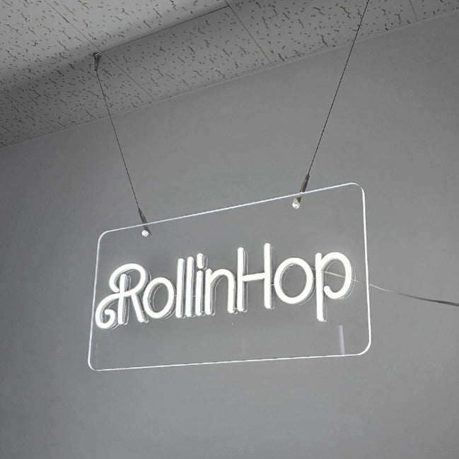 RollinHopロゴのネオン看板（斜め視点）。側面構造と上部から出る電源コード仕様を確認でき、天井設置に対応した仕様。